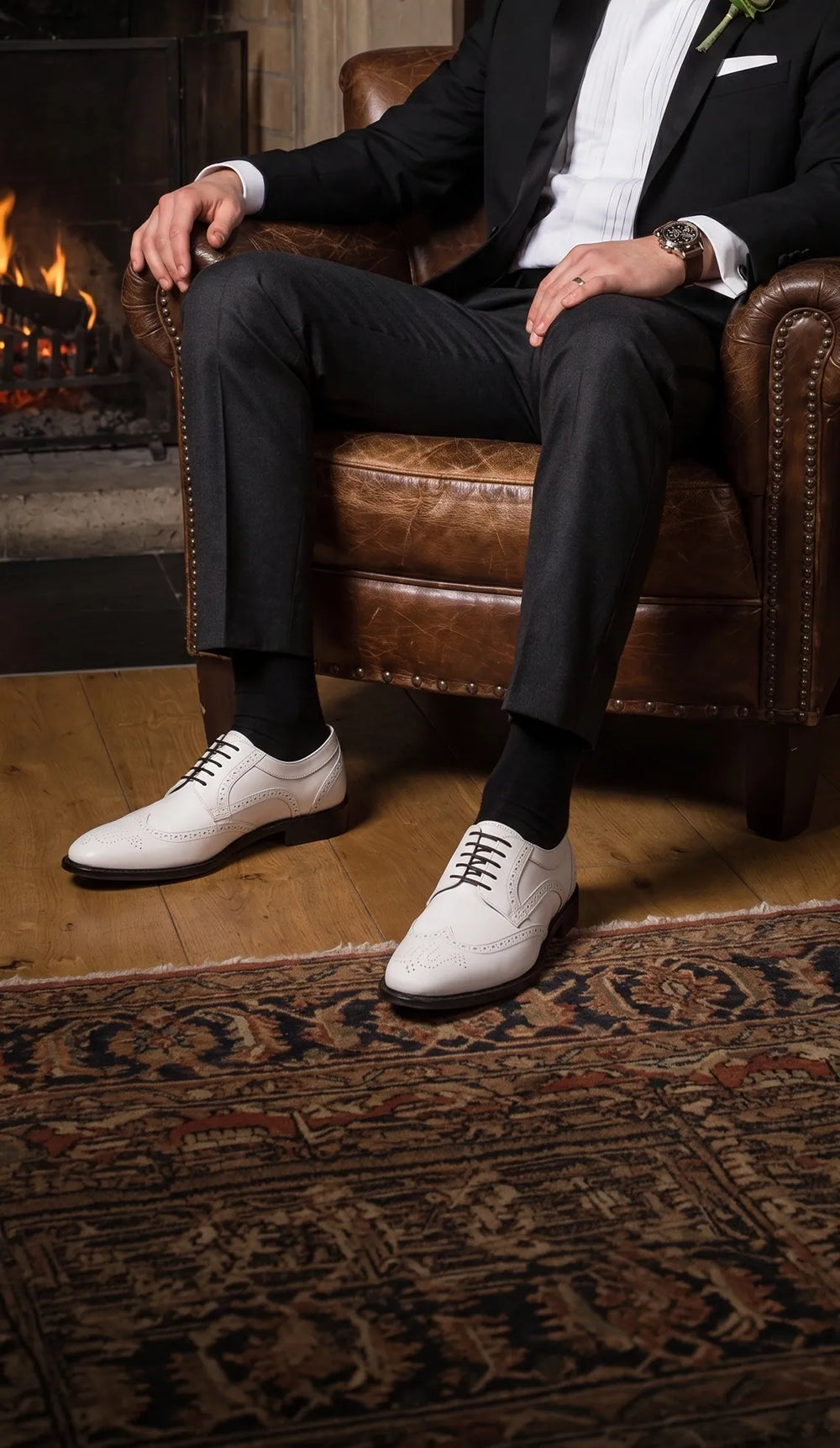 Man in a black tuxedo wearing white and black two-tone brogue dress shoes beside a fireplace